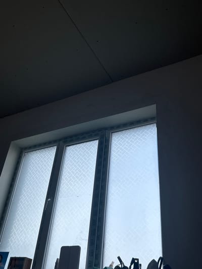 The image shows an interior view of a room in Dmitrievskaya, Russia. The focus is on a window, likely a skylight or attic window due to the sloped wall and ceiling. The window is white-framed and appears to be divided into multiple panes. The glass has a textured or patterned covering, possibly for privacy or light diffusion. To the left of the window, the wall is unfinished, with visible drywall and uneven surfaces. The overall lighting is dim, with the window providing the main source of light.