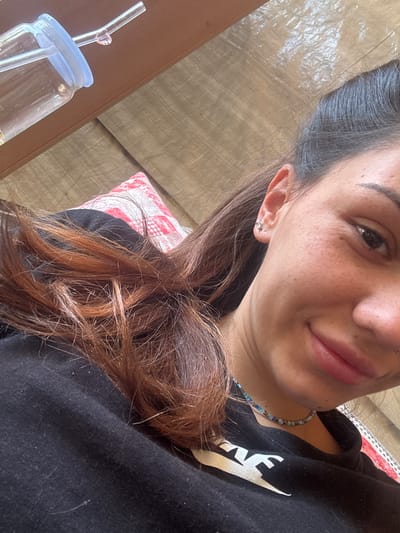 In Belopoptsi, Bulgaria, a close-up selfie shows a woman's face and upper body. She is wearing a black t-shirt with a white graphic and a beaded necklace. Her hair is dark brown with auburn highlights, and she has a small stud earring in her left ear. Her skin has a visible texture with freckles. In the background, a glass cup with a straw is visible, resting on a wooden surface. A patterned pillow is also partially visible behind her. The lighting suggests an indoor setting, possibly daytime.
