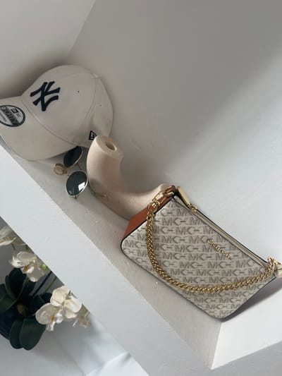 The scene, located in Skhirat, Morocco, depicts several personal accessories arranged on a white, smooth surface, likely a shelf or ledge, with a contrasting diagonal architectural element visible in the lower left. On the left, a white baseball cap, identified by a "NY" logo in dark blue and a "9FORTY ADJUSTABLE" tag, rests. Adjacent to the cap are a pair of gold-framed sunglasses with dark, circular lenses. To their right, a light beige, curved, and hollow object with a circular opening at one end is positioned. On the far right, a Michael Kors brand clutch or small shoulder bag is visible. The bag features a repeating light grey "MK" monogram pattern on a lighter background, brown leather accents on its sides, and a prominent gold-colored chain strap. The "MICHAEL KORS" brand name is visible on the bag's exterior. Partially visible in the bottom-left corner, beneath the white diagonal structure, are a few white orchid flowers with green leaves, emerging from a dark container. No people are present.