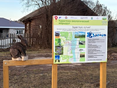 Here is a detailed description of the image:

The image presents a rural scene in Дулесово, Russia, centered around a wooden signpost. The sign, titled "Tourist-Ecological Route," is in Russian and provides information about the "Дулесовскими Тропами" (Dulesovo Trails) project, scheduled for 2025. It includes an invitation to explore the ecology, history, archaeology, and literature of the area, along with a character called "Дулесовский Валенок" (Dulesovo Valenok). The sign includes a map of the area, highlighting attractions such as the Shitoletovo Bridge, Fox Mountain, and the Kama River, among others. It also advertises events like the "Valenok Holiday" and excursions along the "Dulesovo Trails," and lists two telephone numbers for more information.

To the left of the sign is a wooden sculpture of a rabbit, resting on a small platform attached to the wooden fence-like structure of the signpost. The background includes a partly visible log cabin with an old, weathered roof and a newer house with a grey roof in the distance. The environment appears to be somewhat muddy or earthy, suggesting a rural or semi-rural setting. The sky is overcast, and there's a hint of foliage, although much of it looks bare, indicating possibly late autumn or early spring.