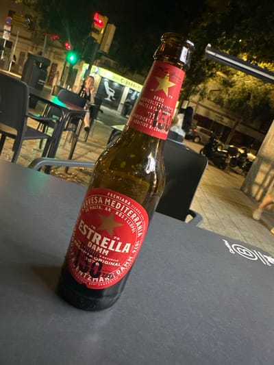 An eye-level, slightly angled medium shot shows a bottle of Estrella Damm beer resting on a dark gray table outside a cafe. The beer bottle, in the foreground and leaning to the right, has a red label with gold text and a gold star. The label reads "CERVESA MEDITERRANEA ESTRELLA DAMM", with additional text that is partially obscured. Behind the table, a woman in a black dress is visible sitting at another table, talking on her phone and smiling. She is seated at a table with chairs that appear to be made of dark metal and plastic. In the background, the street scene of Palma, Spain, is illuminated by streetlights and shop signs. A traffic light is visible with green and red lights illuminated. Storefronts with closed roll-up shutters are present, including one that says "BARBER". Several motorcycles are parked on the sidewalk. The overall atmosphere suggests a casual evening at an outdoor cafe in a lively urban environment.