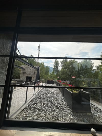This image captures an outdoor landscape and architectural elements viewed through a large, dark-framed window, likely located in Aspen, United States.
The foreground, seen from inside, reveals a segment of a light-toned wooden floor at the bottom. A small, white spherical object, possibly a smart speaker, rests on this floor in the lower right.
Outside the window, a meticulously landscaped area is visible. To the left, a grey stone structure with an integrated outdoor fireplace stands adjacent to a grey composite or planked patio. A dark, low-level railing borders this patio. The central ground is entirely covered with a dense layer of light grey, smooth, rounded river pebbles. Parallel to the pebble area on the right is a long, dark-colored planter box containing a variety of green plants, vibrant red flowers (possibly geraniums), and purple spiky flowers. Two red hummingbird feeders are suspended above the planter on dark hooks, and a portion of an American flag is visible within the planter towards the far right.
The background consists of a dense treeline dominated by various green trees, extending towards distant mountains shrouded in a light haze. The sky is partially cloudy. Prominently positioned on the left side of the background, rising above the trees, is a tall, dark, multi-segmented mast or boom, consistent with a construction crane or telecommunications tower. No individuals or direct human actions are observed within the scene.