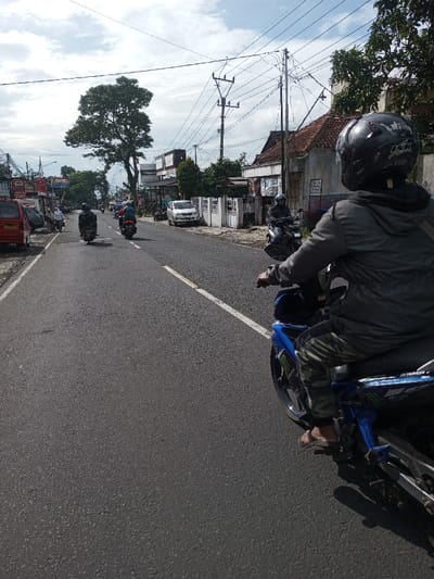 The scene depicts a daytime street in Domphoog_Dungkarchhoeling, Bhutan, viewed from the perspective of a person on a motorcycle. The asphalt road features a solid white lane divider that transitions to a broken line further down.
In the immediate foreground, facing away from the viewer, a person is seated on a blue Yamaha motorcycle. This individual wears a black full-face helmet with white markings, a dark grey hooded jacket, camouflage-patterned trousers, and dark sandals. The motorcycle exhibits blue bodywork with dark green/black graphic decals. A shadow from the foreground motorcycle is cast onto the road surface.
Ahead, three other motorcycles are visible, traveling away from the camera in the same direction. Their riders are also helmeted and clad in dark clothing. On the far left of the road, a red minivan or small car is parked or moving slowly, followed by additional parked vehicles and at least one pedestrian, dressed in white, walking on a sidewalk or shoulder.
The background along the left side of the street features multiple buildings, some displaying commercial signage with partially visible text, including "RKI." A large, mature tree with dense green foliage stands prominently on this side. Along the right side of the street, a white building with a reddish-brown tiled roof is visible, along with white fences featuring decorative metalwork. Numerous utility poles with a complex network of overhead power lines crisscross the sky, particularly on the right side. The sky is overcast with significant cloud cover but indicates a bright daytime setting.