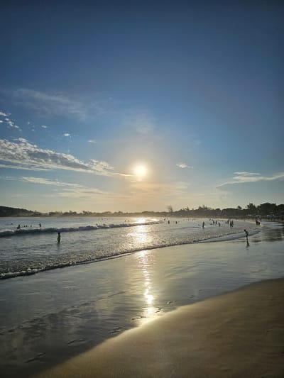 The image captures a beach scene in Armação dos Búzios, Brazil, likely during sunset. The sun is positioned near the horizon, partially obscured by clouds, casting a bright glare across the water's surface. The sky is predominantly blue, with wisps of white clouds scattered. Along the shoreline, people are visible, some standing on the sand, while others are wading in the water. The beach itself appears to be sandy, with a mix of wet and dry areas, reflecting the sunlight. The shoreline is lined with trees and buildings.