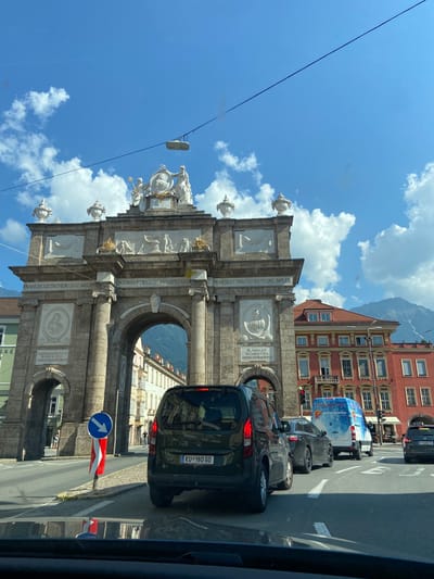 The image captures a street scene in Innsbruck, Austria, likely near the Triumphal Arch. In the foreground, the rear of a dark green van with a license plate reading "KU-180 GD" is visible. Traffic is moderate, with other vehicles including a black car and a white van with a blue and white advertisement on its side. A red and white striped traffic cone with a blue and white directional arrow sign is positioned on the right side of the road. Behind the traffic, a grand archway, part of the Triumphal Arch, dominates the central part of the image. The arch is adorned with intricate sculptures and inscriptions. To the right of the arch, a red building with multiple windows and ornate architectural details is prominent. In the background, a mountain peak, partially covered by snow, rises against a clear blue sky with scattered white clouds. The sun is shining, casting shadows on the street. There are a few pedestrians visible on the sidewalk in the distance near the arch.
