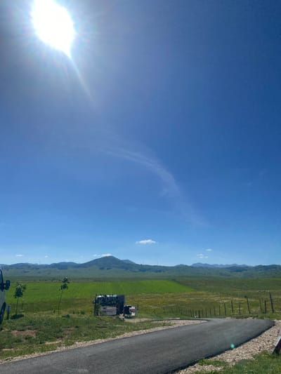 A bright sun shines in the upper left corner of a clear blue sky over a rural landscape. In the middle ground, a field of green grass is dotted with yellow flowers and bisected by a wooden fence. A winding asphalt road occupies the foreground, curving to the right and leading towards a distant mountainous horizon. To the left of the road, a white vehicle is partially visible, and a few small trees are planted in the grassy verge. A small structure, possibly a shed or trailer, is overturned on the grass to the right of the trees. The scene is located in the city of Javorje, Montenegro.