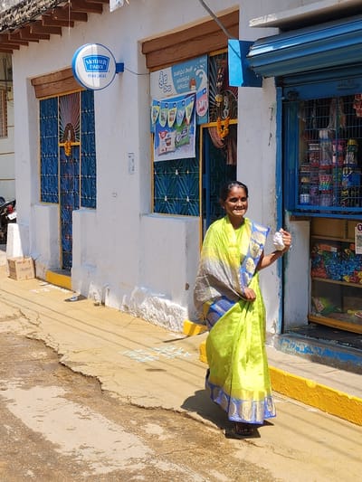 An adult female stands on a concrete path in front of a white-painted building in Pedda Kammavaripalli, India, under direct sunlight. She is positioned slightly to the right of the frame's center, facing the viewer with a smile. She wears a lime green sari with blue and gold borders and dark sandals, and has a red bindi on her forehead. Her right hand holds a small white, crumpled object.
The building behind her features a textured white facade with a yellow painted base. To the left, a blue-framed doorway with an intricate metal grille is adorned with an orange flower garland. Above this, a circular blue and white "MOTHER DAIRY maa jaisi" sign is mounted. Further left, a partial view of a parked dark-colored motorcycle is visible next to a brown cardboard box labeled "Rocket." To the right of the woman, a storefront is visible with a partially raised blue roll-down shutter, revealing shelves stocked with numerous packaged items. A QR code sticker is affixed inside the storefront. A second blue-framed window/doorway with a grille is present to the left of the storefront. A poster with text in a local language (likely Telugu) is affixed to the wall between the two doorways. The path shows cracks and discoloration, with white chalk-like patterns on the ground near the building. Shadows are cast by the woman and the building, indicating bright overhead sunlight.