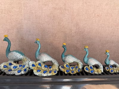 Six decorative peacock figurines are arranged in a line on a dark, ribbed surface against a textured, light brown wall. The peacocks vary in size, decreasing from left to right. Each figurine features a teal body with silver accents, a golden crown, and a tail fanned out with gold and blue feather-like details. The background wall appears to be a textile or suede-like material with a subtle, uneven texture. The scene is lit from the front, casting minimal shadows. The image is taken from a slightly elevated perspective, looking down at the figurines. The location is identified as İzmit, Turkey.