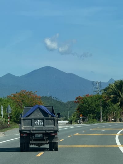 The image shows a dark-colored truck from behind, driving on a paved road in Thôn Thượng, Vietnam. The truck has a blue tarp covering its cargo. The license plate on the truck reads "79A 77136". The road is marked with yellow lines. In the background, there are lush green trees, some with red flowers, and distant blue mountains. The sky is a clear blue with a few white clouds. A utility pole with electrical wires is visible on the right side of the road. A road sign with a triangle and the number "150" is also present. The overall scene depicts a daytime view of a rural or semi-rural road in Vietnam.