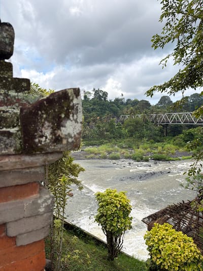 A rustic, partially constructed stone and brick structure stands in the foreground, suggesting a construction site or an unfinished building. Beyond this, a fast-flowing river cuts through a lush, green valley. A prominent metal truss bridge spans the river in the mid-ground, its structure clearly visible against the dense foliage. The riverbanks are lined with vegetation, including trees and bushes, some of which are brightly colored with yellow and green leaves. In the far distance, rolling hills covered in trees create a layered landscape. The sky above is cloudy and overcast, indicating a potentially damp or moody atmosphere. The overall scene conveys a sense of natural beauty intertwined with human infrastructure, possibly in a tropical or subtropical region given the dense greenery. The image appears to be taken during the daytime, with diffused light suggesting cloudy conditions.