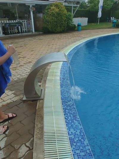 The image shows a person's legs and feet as they stand by a swimming pool. The person is wearing black and red "MARVEL" inscribed sandals and is holding a bottle of water and a blue towel. The swimming pool is filled with blue water and has a tiled border. A water feature is visible, with water splashing into the pool. The area around the pool is paved with interlocking stones and a green lawn with trees is visible in the background. There is a white fence and some outdoor furniture visible. The image appears to have been taken during the daytime, with overcast or mild weather conditions. The location is identified as Yagoda, Bulgaria.