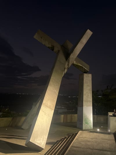The image depicts the "Cruz Caída" (Fallen Cross) monument at Praça da Sé in Salvador, Bahia, Brazil, at night. The monument consists of three large, rectilinear concrete beams. Two elongated beams intersect and lean dynamically, while a third shorter, vertical beam serves as a base on the right. The concrete surfaces display weathering, including dark streaks and discoloration, with green graffiti visible on the lower section of the vertical base on the right.
Artificial ground lighting illuminates the monument, casting distinct, elongated shadows on the concrete platform beneath. In the background, beyond a low wall or railing, the dark expanse of the All Saints Bay (Baía de Todos os Santos) is visible, illuminated by numerous distant city lights on the opposite shore. The sky above is a dark, cloudy blue-black. On the far lower right, a dark, indistinct figure is partially visible, seated on a low, block-like structure. Concrete steps or tiered elements are present in the lower right foreground.