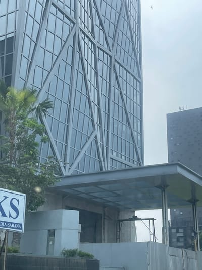 The image shows a modern office building in the city of Medan, Indonesia. The building features a unique geometric facade with a lattice of grey metal beams intersecting a grid of glass windows. In the foreground, there is a smaller structure with a translucent roof supported by grey beams and columns, possibly an entrance canopy or an extended walkway. Construction materials and scaffolding are visible in the area below the canopy, suggesting ongoing development or renovation. To the left, a sign with blue lettering on a white background is partially visible, displaying the letters "KS" and "UMA SARANA" below. Lush green trees and palm fronds are present on the left side of the frame, adding a touch of nature to the urban landscape. In the background, another dark-colored building with a regular pattern of windows is visible. The sky is overcast, contributing to a muted color palette of the scene.