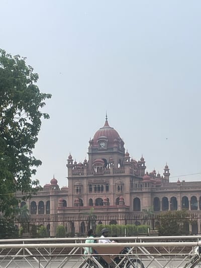 The image shows the Khalsa College in Amritsar, India, on an overcast day. The architectural style is evident, with a prominent dome and intricate detailing on the building's facade. A tree is partially visible in the foreground on the left, its leaves obscuring some of the building's left side. A metal fence separates the building from the foreground. In the lower part of the image, a white, elevated structure, possibly a pedestrian bridge or overpass, is visible, with several people and bicycles in the background. The sky is a light grey, indicating cloudy weather.