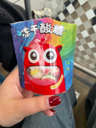 A person holds a colorful bag of candy with a cartoon monster design. The bag features Chinese characters and the text "FREEZEDRY". The candy inside appears to be a mix of round, colorful sweets. The background shows a checkered pattern and a person's lap dressed in denim. The image was taken in Yakovlevskoye, Russia.