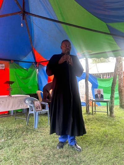 An outdoor gathering is depicted, likely in Kiganjo, Kenya, under a multi-colored canopy with sections of blue, green, and orange/red fabric. The ground is covered in grass, and palm trees are visible in the bright background, indicating clear, sunny weather.

The central subject is a man standing, appearing to be in clerical attire, including a black cassock or robe, blue trousers, and black shoes. He holds his hands together near his chest, possibly holding a microphone or prayer beads, and looks directly towards the viewer. Sunlight streams down from the top right, creating noticeable rays of light.

To the man's right, a light blue plastic chair holds a small framed picture, possibly a religious icon like the Madonna and Child. Further back, another plastic chair and a table draped with a white cloth are visible. Partial text on the cloth reads "PETER S.C.C," suggesting a connection to a St. Peter's Small Christian Community or similar group.

In the mid-ground, to the man's left, a stand displays a framed portrait of another man in a suit, with text "Baby @83" next to his image, possibly indicating a name or age. Other unidentifiable structures are in the background. The scene suggests a community event or religious service taking place outdoors.