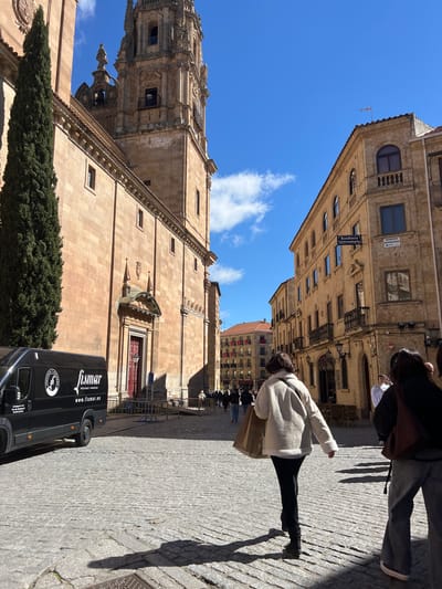 The image depicts a street scene in Salamanca, Spain, likely near the cathedral. The focus is on the architectural detail of the cathedral and the surrounding buildings, with a bright blue sky overhead. A person in a light-colored jacket and dark pants is visible walking away from the camera. A black van with the logo "fismar" is parked on the cobblestone street in front of the cathedral. The environment is sunny and suggests an urban setting within a historic district. Other pedestrians are visible in the background.