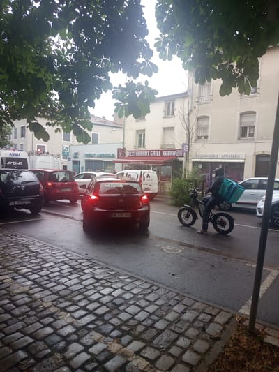 This is a vertical, slightly blurry photograph taken from a low angle, looking up towards buildings and down a street. The dominant elements are a delivery person on an electric scooter and traffic. The delivery person, wearing a blue thermal bag on their back and a helmet, is on a black electric scooter. They are facing towards the right side of the frame, and their scooter is in motion. Behind them, several cars are stopped in traffic, including a red car with its brake lights on, a smaller red car, and a black car with visible license plate text. Further down the street, there are more vehicles and buildings.
The scene appears to be urban, with storefronts visible on the left side of the street. Text visible on these storefronts includes "RESTAURANT GRILL KEBAB," "BOULANGERIE PATISSERIE," and "CREDIT AGRICOLE." A white van with "ASSU DMS" written on it is also present. The street surface is wet, suggesting it might have recently rained or is raining, and the sidewalk is paved with cobblestones. Lush green tree leaves frame the top left of the image, obscuring a portion of the sky and buildings. The lighting suggests it could be daytime, possibly overcast. The overall atmosphere is that of a busy street with ongoing delivery services and traffic.