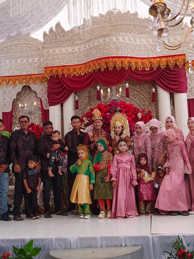 In Meuliek, Indonesia, a wedding ceremony is taking place on an elevated stage, elaborately decorated with white ornate carvings and deep red velvet drapes adorned with gold trim. A large floral arrangement of red roses forms the backdrop for the wedding party. The bride and groom, dressed in traditional wedding attire, stand center. The bride wears a golden headdress and a deep red and gold outfit, while the groom is in a similar red and gold ensemble. They are surrounded by family members and children, all dressed in festive clothing. Men on the left wear matching dark batik shirts, some holding children. Women on the right are mostly in pink hijabs and matching dresses, with several children in colorful traditional garments, including one in a green and gold outfit giving a thumbs-up. The atmosphere appears to be joyful and celebratory, with various decorative elements, including chandeliers, visible in the background.