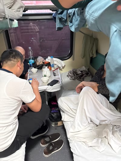 The image depicts the interior of a train cabin. Two people are in the lower berth, one lying down and looking at a phone. A second person is sitting and also looking at the phone. Various personal items are on a small table, including a tea set, water bottles, and a smartphone. The berth above appears to have clothes and blankets on it, and someone is sleeping on the upper berth. The scene suggests travel, and there is a window behind the seated person. The location is unknown.