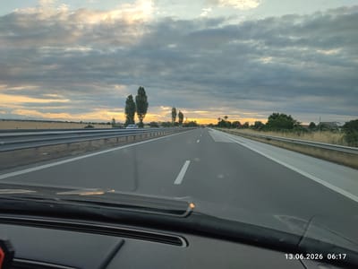 The image shows a perspective from inside a vehicle, looking forward along a highway. The highway has multiple lanes and is bordered by a metal guardrail and dry vegetation. In the distance, the sky is filled with dramatic, overcast clouds with patches of sunlight breaking through near the horizon, suggesting either dawn or dusk. Several tall, slender trees are visible along the side of the road. There are some distant buildings and fields to the left, and more trees and buildings to the right. The dashboard of the vehicle is partially visible in the foreground, with a timestamp in the lower right corner reading "13.06.2026 06:17". The scene appears to be a highway near Tsaratsovo, Bulgaria, given the date and time stamp's proximity to local sunrise. No individuals are directly visible, and the focus is on the road and the surrounding landscape under a dynamic sky.