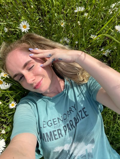 The image shows a woman lying on her back in a field of daisies and grass. She has blonde hair and is wearing a teal t-shirt with text on it. Her left hand is raised to her forehead, shielding her eyes from the sun. The background is filled with green grass and white daisies. There is no visible indication of the city of Yasnoe, Russia, or any other specific location.