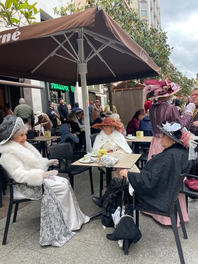 The image shows a group of elderly women dressed in period costumes, sitting at tables outside a cafe in Terrassa, Spain. The women are wearing elaborate hats and dresses. Some of the women are interacting with each other. There are other people at the tables that are not in costume. The cafe has brown umbrellas with the word "Carne" printed on them. There are trees and buildings in the background. The weather appears overcast.