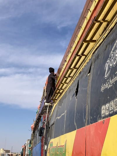 A male individual in a black beanie and dark shalwar kameez is observed ascending a metal ladder attached to the side of a large, multi-colored vehicle, likely a passenger bus or cargo truck, in Chaman City Tehsil, Pakistan. The individual is positioned on the ladder, reaching towards the vehicle's upper structure. The vehicle's side is predominantly black, featuring white text in Arabic script and several white icons (e.g., airplane, Wi-Fi, no smoking, air conditioning). Below the black section are horizontal yellow and red panels. The upper edge of the vehicle displays a yellow and red frame with multiple horizontal bars and attachment hooks. Visible cracks and a tear are present in the black side paneling. Items, possibly cargo, secured with orange straps are visible on the vehicle's roof. Other vehicles and distant buildings are faintly visible on the left side of the frame. The sky above is clear blue with sparse white clouds, indicating daytime.