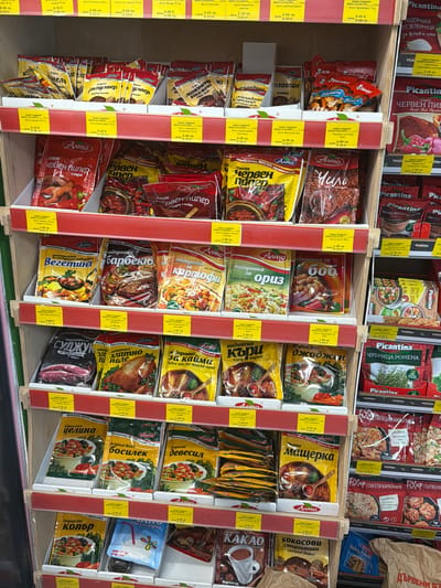 The image shows a retail display of various food seasonings and spices in a store. The shelves are organized with products in colorful packaging, predominantly featuring red, yellow, and green hues. The products are arranged in rows and sections, with some items displayed in clear plastic containers or small baskets. Labels on the packaging are in Bulgarian, indicating brand names like "Arno" and product types such as "червен пипер" (red pepper), "чубрица" (savory), and "босилек" (basil). Some products have yellow price tags or informational stickers with pricing and product details.
The background of the image is a light-colored wall or shelving unit, with some wooden elements visible in the structure of the shelves. The lighting appears to be from overhead fixtures, casting a consistent illumination across the products. The perspective is slightly angled, looking up at the shelves.
The retail setting suggests a supermarket or a specialty food store. While the image does not explicitly show any people, the presence of price tags and organized displays implies commercial activity. The specific location mentioned, Ravno Pole, Bulgaria, places this scene within a Bulgarian context, where Bulgarian language product labeling would be expected.