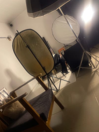 The image depicts an indoor studio setup, likely for photography. In the foreground, a wooden chair with a dark cushioned seat is visible, with a fluffy, light-colored object, possibly a pet, resting on the cushion. To the left and slightly behind the chair, a large, oval-shaped photography reflector with a gold-colored side is propped up by a stand. Further into the background, to the right, a person is seated on a chair, partially obscured, wearing dark clothing. In the upper right quadrant of the image, a bright studio light illuminates a white softbox or diffuser. Another large black backdrop on a stand is positioned to the right of the person. The environment appears to be a room with tiled flooring and white walls, suggestive of a studio space. There is no indication of the location being in South Ropp, Nigeria.