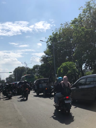 The image depicts a traffic jam on a sunny day on a street in Banjarkemantren, Indonesia. A variety of vehicles are present, including motorcycles, cars, and a bus. On a motorcycle in the foreground, a person wearing a blue helmet and a pink headscarf is riding as a passenger. The motorcycle has a license plate with the registration "W 5633 NGO". In the background, a green bus is visible, and further down the road, traffic lights are illuminated green. Lush green trees line the right side of the street, and the sky is bright blue with scattered white clouds. The overall scene captures a moment of congested urban traffic.