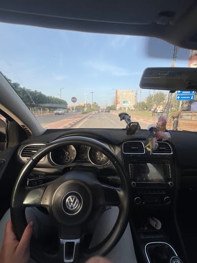 The image shows a view from inside a Volkswagen car, specifically the steering wheel and dashboard, facing forward. The car is driving on a street in Beni Mellal, Morocco. Visible outside the windshield are buildings, trees, street signs, and other vehicles. A street sign indicates directions to Khouribga and Casablanca. Construction appears to be taking place on a nearby building. A decorative object hangs from the rearview mirror. A hand is visible on the steering wheel.