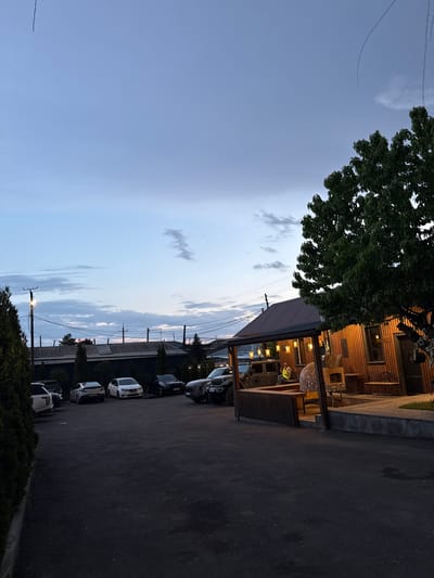 The image shows a street scene in Arinj, Armenia, likely at dusk or evening. The sky transitions from a light blue to a darker shade. A line of parked cars, mostly light-colored SUVs, runs along the right side of the street. Overhead power lines are visible, supported by utility poles. A building with corrugated metal siding and a covered porch sits on the left. The porch area is lit with string lights and contains furniture. A person wearing a bright yellow vest is visible on the porch. A large tree partially obscures the building. The road appears to be asphalt.
