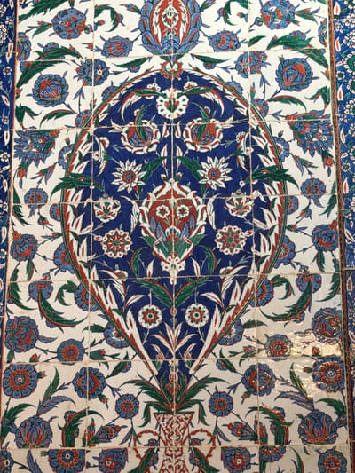 The image shows a close-up of a tiled wall with a floral pattern. The tiles are square and arranged in a grid. The pattern features blue and red flowers with green leaves on a white background. The main design in the center is an oval shape with elaborate floral motifs in blue, red, and white. The overall style is reminiscent of Iznik pottery. This type of tile work is common in historical buildings in Eyüpsultan, Istanbul, Turkey, particularly in mosques and tombs.