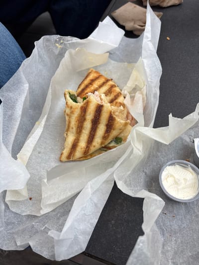 A partially consumed panini-style sandwich, cut into two halves, rests on crumpled white wax paper. The sandwich displays prominent grill marks on its bread, revealing fillings of light-colored deli meat and green leafy vegetables. A bite has been taken from one of the halves. To the right, a small, gray plastic container holds a white, creamy substance with visible swirls, consistent with cream cheese. The food items are situated on a dark, textured surface, likely a table. A portion of a person's leg, clad in blue denim jeans, is visible in the upper left. A crumpled brown paper bag is partially visible in the upper right background. Small food debris is scattered on the wax paper and table. The specific location within the city of Wellesley, United States, is not discernable from the image.