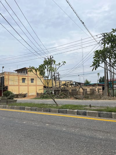 In Tanahterban, Indonesia, an image captures a street scene with a damaged building and overhead power lines under a cloudy sky. The building on the right side of the image appears to be undergoing demolition or has been partially destroyed, with its roof structure exposed and debris scattered around its base. To the left, a yellow multi-story building stands intact, featuring windows and visible structural beams. The foreground shows an asphalt road with a yellow dividing line and a curb with alternating black and grey stones, bordered by a strip of grass. Trees are visible throughout the scene, including one in the mid-ground with fallen branches and leaves on the ground, and others with green foliage to the right. Numerous power lines crisscross the sky above the buildings and road, supported by utility poles. In the background, a tall communication tower is visible. No people are present in the image.