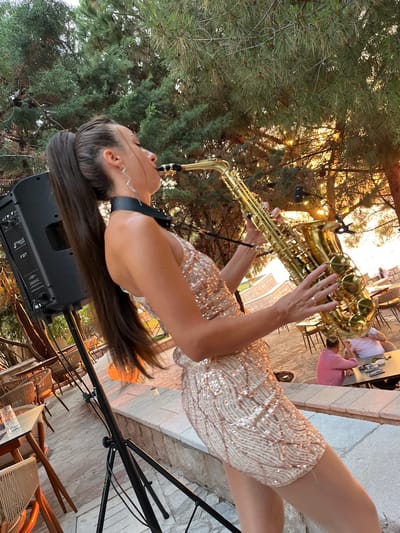 A woman with long dark hair in a high ponytail plays a gold saxophone outdoors in front of a black speaker. She is wearing a sparkly, short, sleeveless dress. Her head is tilted up, and her eyes are closed as she plays. The background shows a sun-drenched outdoor seating area with tables and chairs, trees, and a glimpse of the sea. There are other patrons seated at tables in the background. The location appears to be a restaurant or bar on the coast.