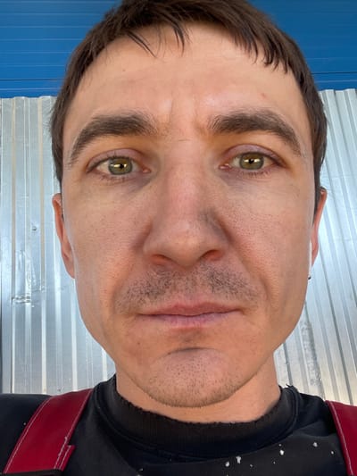 The image is a close-up, front-facing selfie of a man with brown hair, green eyes, and light stubble on his face. He is wearing a black t-shirt with red suspenders. The background consists of a corrugated metal wall and a blue surface. The lighting is natural, casting shadows on the man's face. The photo appears to have been taken outdoors, possibly in a rural or industrial area. The man's expression is neutral. No specific location in Sokury, Russia, can be identified from the visual information provided.