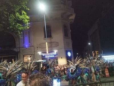 A night scene captures a vibrant street performance. Elaborately costumed individuals with feathered headdresses and painted faces are in the foreground. A crowd lines the street behind them. A Starbucks Coffee shop is visible, brightly lit with its sign. Above, a building with architectural details and arched openings adds to the urban backdrop. A streetlight shines intensely in the upper center.
