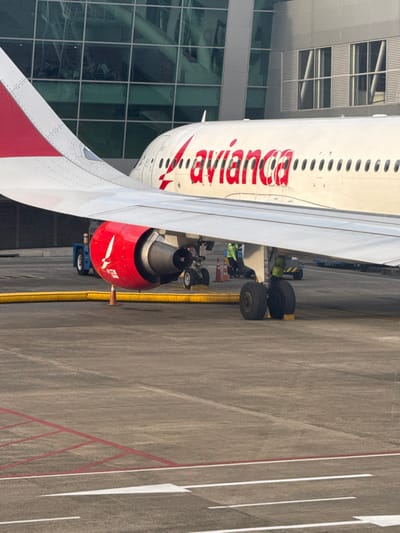 An Avianca aircraft is parked on an airport tarmac. The plane's fuselage, painted white with red lettering, is visible along with a wing featuring a red tip. A red jet engine is positioned under the wing. The plane's landing gear is visible near the fuselage. A modern airport terminal with a glass facade stands in the background. The tarmac has painted lines and safety markings.