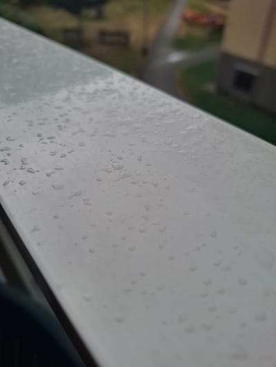 This is a close-up, slightly angled shot of a white railing covered in numerous water droplets. The background is blurred, showing a green grassy area, a road, and a building with a yellow facade and dark windows. The scene appears to be outdoors, possibly on a balcony or railing overlooking a street. The focus is on the textured surface of the railing with the water droplets.