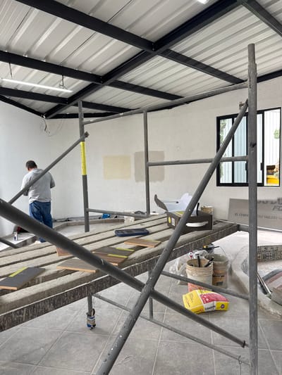 A construction scene inside a building in San Francisco Totimehuacán, Mexico is depicted. A metal scaffold dominates the foreground. A man in a gray shirt and blue jeans stands on the scaffold. Construction materials (buckets, a bag of mortar) are on the scaffold. The building has white walls, a window, and a corrugated metal ceiling with black metal support beams. A light tube is affixed to the support beams. Tiles are on the floor in the background.