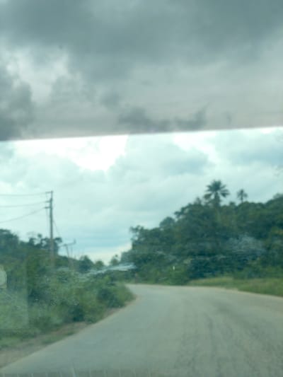 The image depicts a paved road curving to the right, bordered by dense green vegetation on the left and a tree-covered hillside on the right. The sky is overcast with heavy, gray clouds. In the background on the left, a utility pole with wires is visible. The overall scene suggests a rural or semi-rural environment, possibly on the outskirts of a settlement like Iyah Gbede, Nigeria. The road appears to be a secondary route, with no other vehicles or people immediately apparent in the frame. The image is taken from a slightly elevated perspective, possibly from inside a moving vehicle, as evidenced by the slight blur and reflections on what appears to be a windshield.