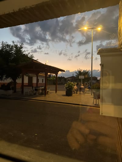 The image captures an outdoor scene in Arcahueja, Spain, at dusk. The sky is filled with dramatic, dark clouds illuminated by the soft orange glow of the setting sun on the horizon. A double-headed street lamp with bright, glowing lights stands prominently in the mid-ground. To the left, a wooden pergola covers a picnic table with chairs, suggesting a public seating area. Behind the pergola, a building with a white and orange facade is visible. In the distance, a basketball hoop and backboard are set up.
On the sidewalk in front of the basketball hoop, three individuals are seated on chairs, appearing to be in conversation. One person is wearing a blue shirt, another a light-colored shirt, and the third a dark shirt. A large potted plant with white flowers sits to the left of the group.
The foreground is dominated by a dark asphalt road with a yellow line marking the edge of the sidewalk. A reflection of what appears to be a person or persons is cast on the lower portion of the image, obscuring details of the road surface. The overall impression is one of a quiet evening in a small town.