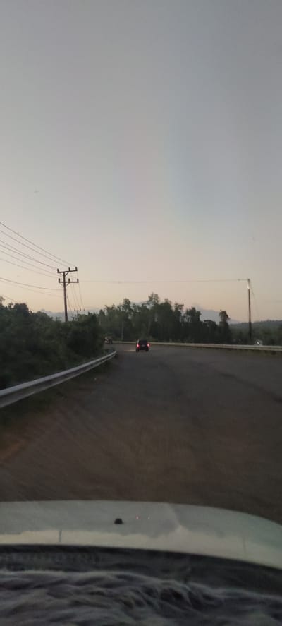 This image depicts a road scene in Kareungateuh, Indonesia. A dark-colored car is visible driving away from the viewer on a paved road. To the left of the road is a silver guardrail, and beyond that, lush green trees and vegetation. Several utility poles with wires are also present in the background, interspersed with the trees. The sky is overcast and a pale gray, suggesting either early morning or late evening. The foreground shows the dashboard and windshield of a vehicle, indicating the photograph was taken from inside a moving car. The overall environment appears to be rural or semi-rural, with a focus on the road and the surrounding natural landscape.