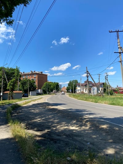 The image shows a curved asphalt road in a semi-urban environment. The road is bordered by a sidewalk on the left and a grassy area on the right. Power lines are visible throughout the scene, supported by concrete and wooden poles. Several buildings, including a multi-story brick apartment building and some single-family homes with decorative roofs, are situated along the road. A white car is seen driving away from the viewer on the road, and a dark-colored car is parked on the side near the apartment building. The sky is bright blue with scattered white clouds. The ground along the side of the road appears to be a mix of dirt, gravel, and overgrown grass. The scene is well-lit, suggesting it is daytime with sunny conditions. The environment depicts a typical residential area with infrastructure for power distribution.