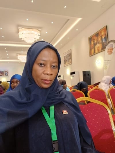 The image captures an indoor scene, likely a conference or event in Unguwan Sanusi, Nigeria. The primary subject is a woman in the foreground, wearing a dark blue hijab and a matching top, looking directly at the camera with a neutral expression. She has a nose piercing, and a bright green lanyard with a black plastic buckle is visible around her neck. A small red and white lapel pin is attached to her attire.

Behind her, another person in a blue hijab is partially visible, along with several other individuals seated in the background. The setting features rows of distinctive red chairs with golden frames. The room's decor includes light-colored walls and ceiling, adorned with decorative, ornate light fixtures resembling modern chandeliers, and framed artwork. White air conditioning units are mounted on the walls. In the far background, a large circular standee with a man's face and some unreadable text is visible. The overall impression is a formal gathering in a well-lit, contemporary event space.