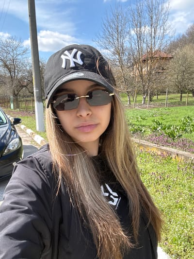 A woman wearing a black New York Yankees baseball cap, black sunglasses, and a black jacket is taking a selfie outdoors. Her long, highlighted hair is visible. Part of a dark-colored car is visible in the bottom left corner of the image. In the background, there is greenery, including grass and purple flowers. Bare trees are present, along with a house partially visible among the trees. The sky is blue with some clouds. While the photo is in Ralevo, Bulgaria, there are no identifiable landmarks that can confirm the specific location. The scene depicts a casual outdoor setting, likely in a rural or suburban area, with the woman as the central subject.