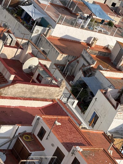 This image captures a complex, densely packed residential building facade, viewed from an elevated, rotated perspective that creates an abstract and disorienting composition. The setting appears to be an urban or coastal residential area, likely in Peníscola, Spain, given the architectural style and sun-drenched appearance.
The buildings feature multiple stories with numerous balconies and terraces, predominantly constructed from concrete and reddish-brown brick, often contrasted with white painted sections. Many balconies are equipped with white metal railings, some fortified with mesh.
Key subjects and objects include:
- **Awnings and Shades:** A prominent blue and white striped awning hangs from a balcony on the left. Other sun-protective elements include a light blue tarpaulin covering a lower area, a white triangular shade sail, and a folded orange umbrella-like structure.
- **Satellite Dishes:** Two white satellite dishes are visible, one large and central, another smaller one towards the upper right.
- **Utility Installations:** Various wires, cables, and antenna poles crisscross the scene. External air conditioning units are mounted on walls, and a yellow coiled hose is visible on a wall in the lower right section.
- **Greenery:** Numerous potted plants and small gardens adorn the balconies and terraces, adding pockets of green amidst the concrete and brick.
- **People:** On a balcony beneath the blue and white striped awning, a person in a white top is partially visible, appearing to be engaged in a routine activity, possibly leaning or tending to something.
The primary activity depicted is everyday residential life. The presence of awnings and shades, along with the bright, direct sunlight, suggests a warm climate. The strong shadows indicate a time of day between mid-morning and late afternoon. The overall impression is one of practical, lived-in spaces in a vibrant, densely populated environment. No visible text is clearly discernible in the image.