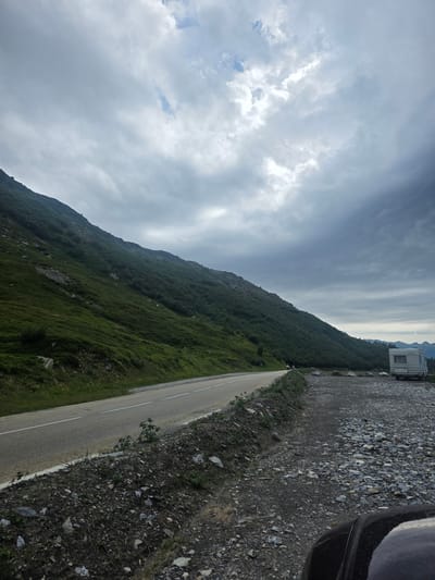 An overcast, cloudy sky looms over a rugged mountain landscape. A paved road with white lines curves along the base of a steep, green, grassy mountain. To the right of the road, a gravel parking area is visible, with a white camper van parked at the far end. The foreground shows the hood of a dark vehicle. The scene is a roadside pull-off, likely in a mountainous region of Séez, France.